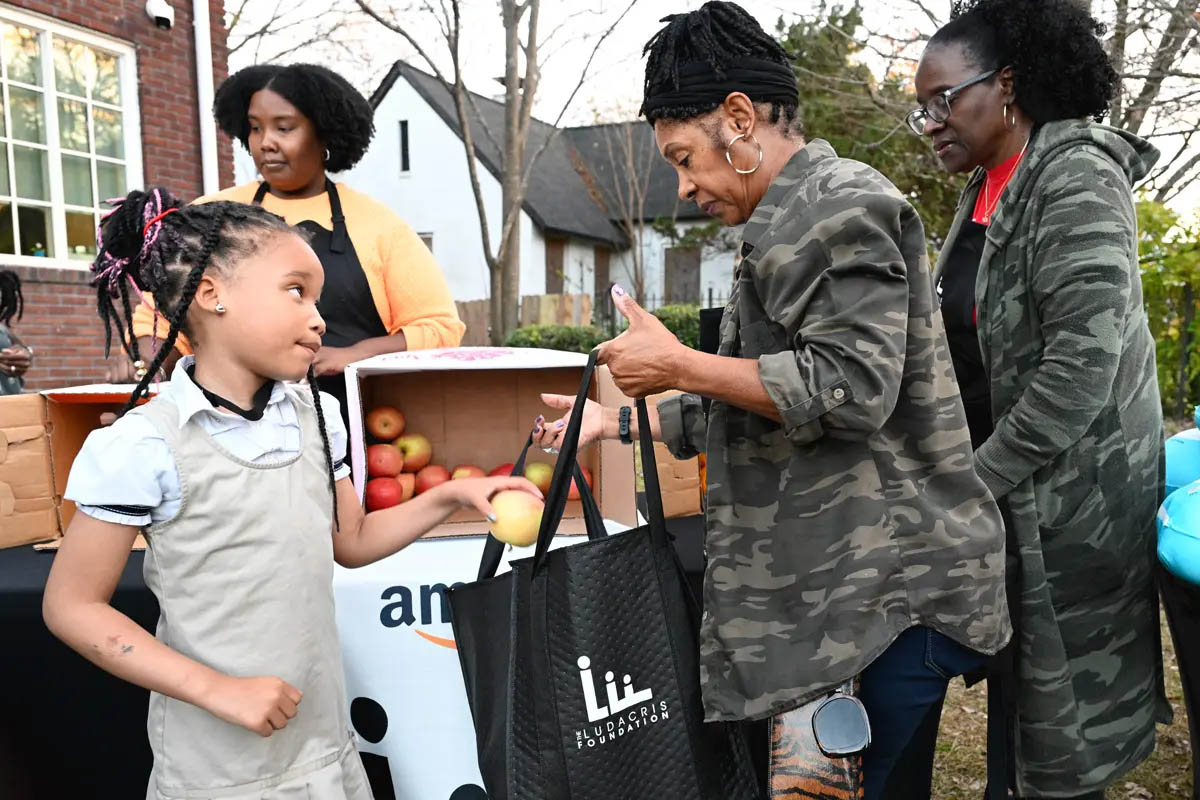 A woman holds a Ludacris Foundation shopping bag as a young girl places an apple in the bag.