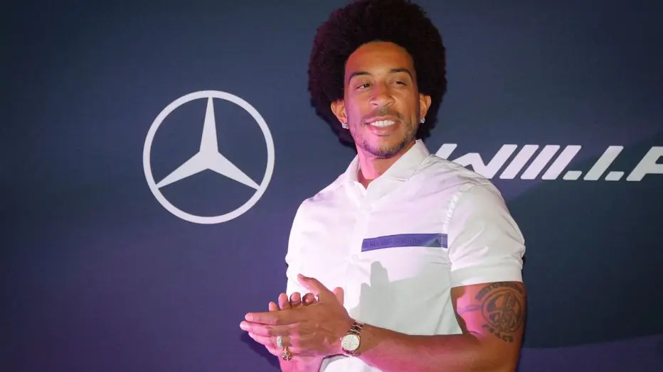 Chris 'Ludacris' Bridges smiles and clasps his hands. He's standing in front of a blue background with the Mercedes-Benz logo.