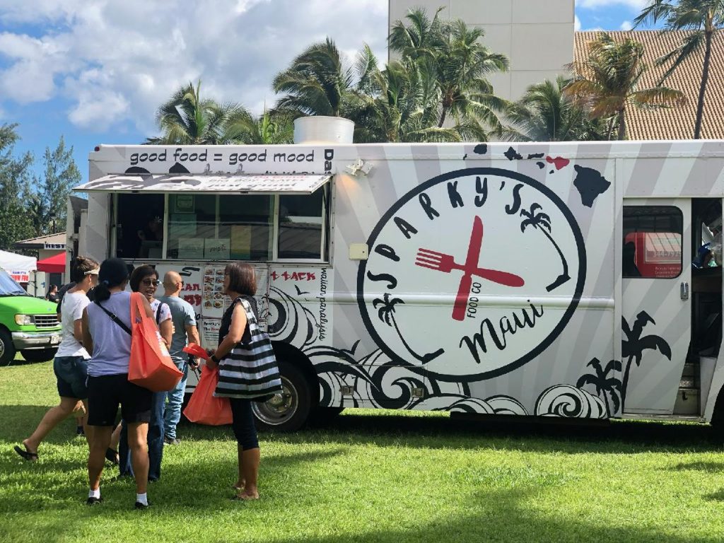 An image of a food truck with the name 'Sparky's Maui' on the side. A line of people is waiting beside the truck.