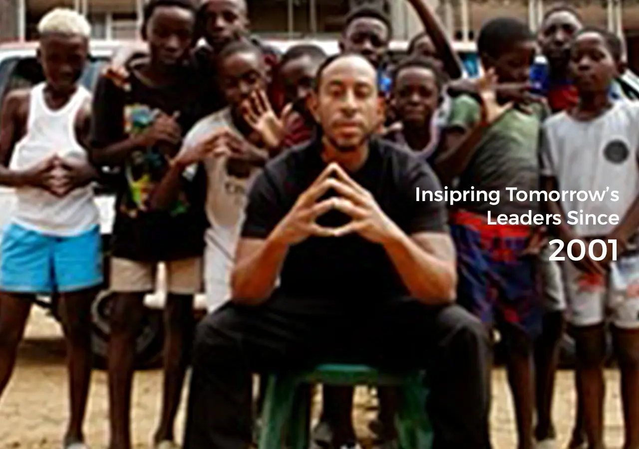 Ludacris stands with a child. A group of people stand in the background. Text reads 'Inspiring Tomorrow's Leaders Since 2001.'