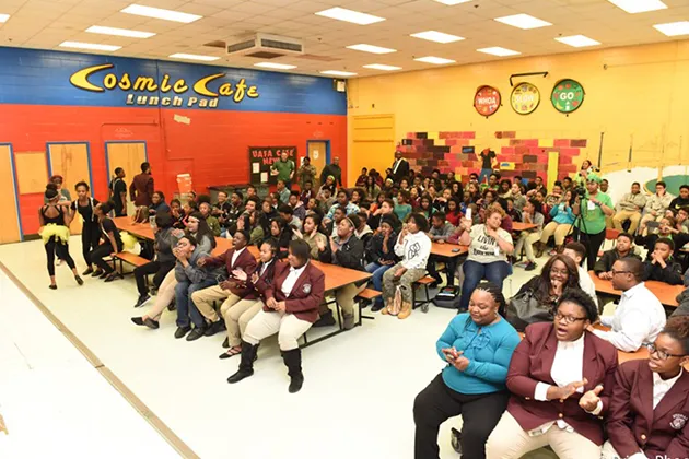 A large group of people sitting at tables in a cafeteria. The cafeteria has 'Cosmic Cafe Lunch Pad' painted on the wall.