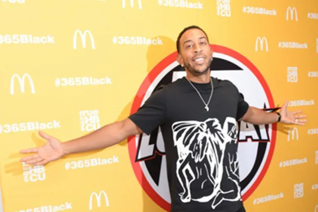 Chris 'Ludacris' Bridges wearing a black t-shirt, standing in front of a yellow background with his arms outstretched.