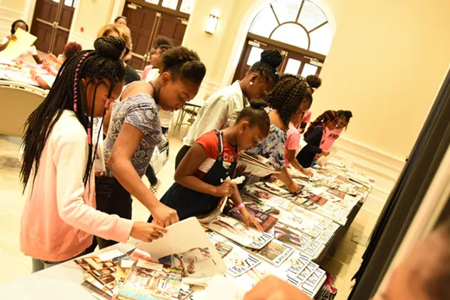 A group of people in a line looking down at various papers and flyers on the tables in front of them.